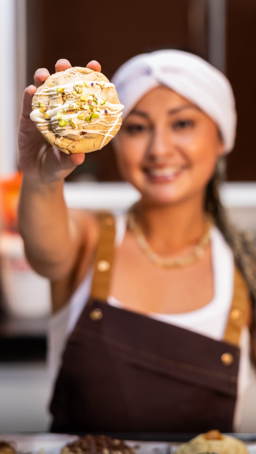 Confeiteira da Magic Cookie, de turbante branco e avental marrom, estende um cookie de pistache para a câmera